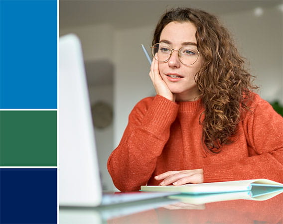 Woman in orange sweater sitting at desk, looking at a laptop, pen in hand
