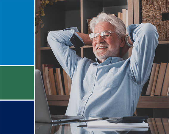 Older man at desk, with his hands behind his head, smiling